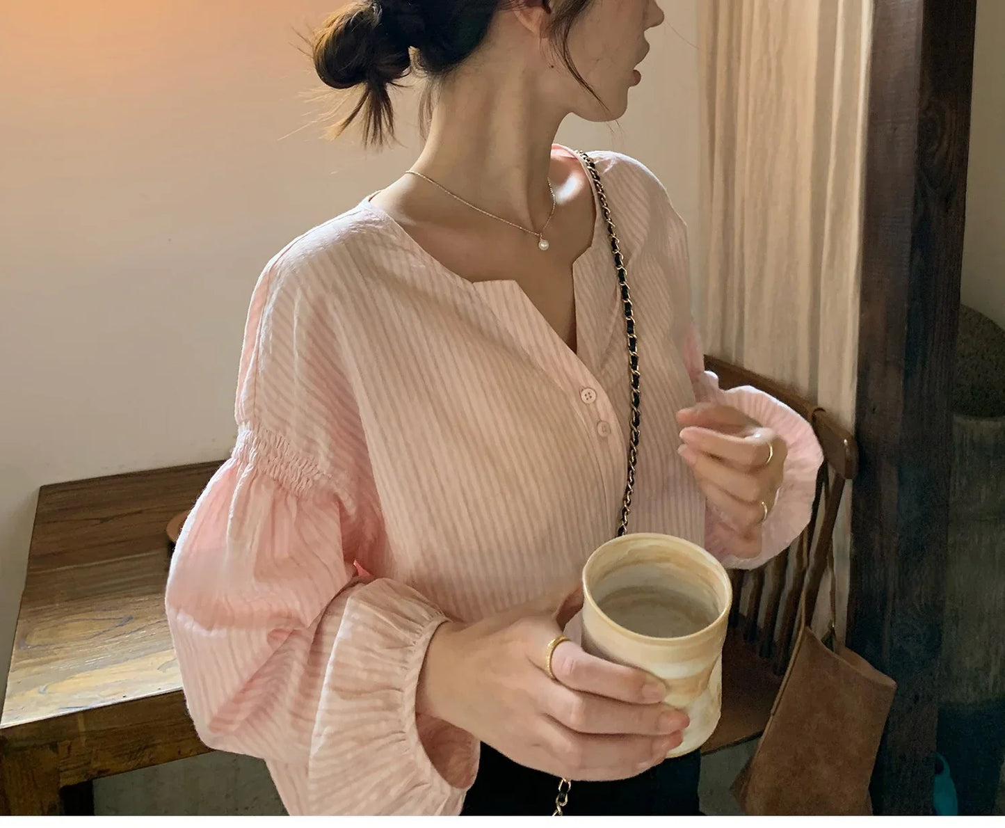 Woman in a light pink blouse with balloon sleeves holding a ceramic cup indoors