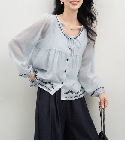 Woman wearing a light blue sheer blouse with black embroidery, long sleeves, and black buttons.