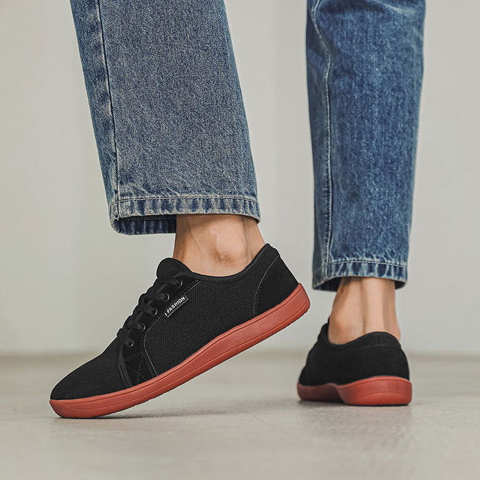 Black sneakers with red soles worn with blue jeans on a light wooden floor.